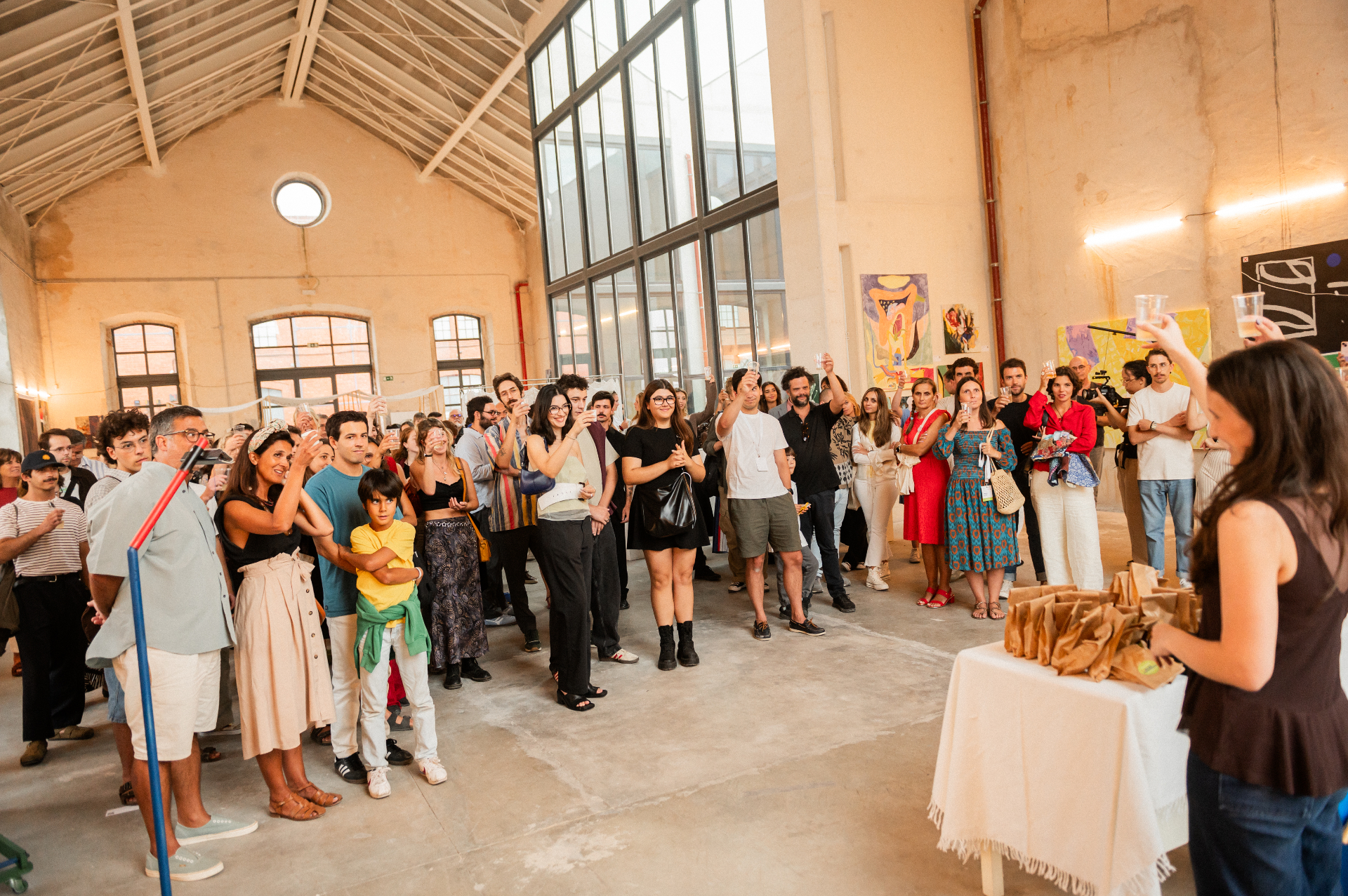 Volume V opening toast at Mitra: crowd raising plastic cups beneath a tall arched window